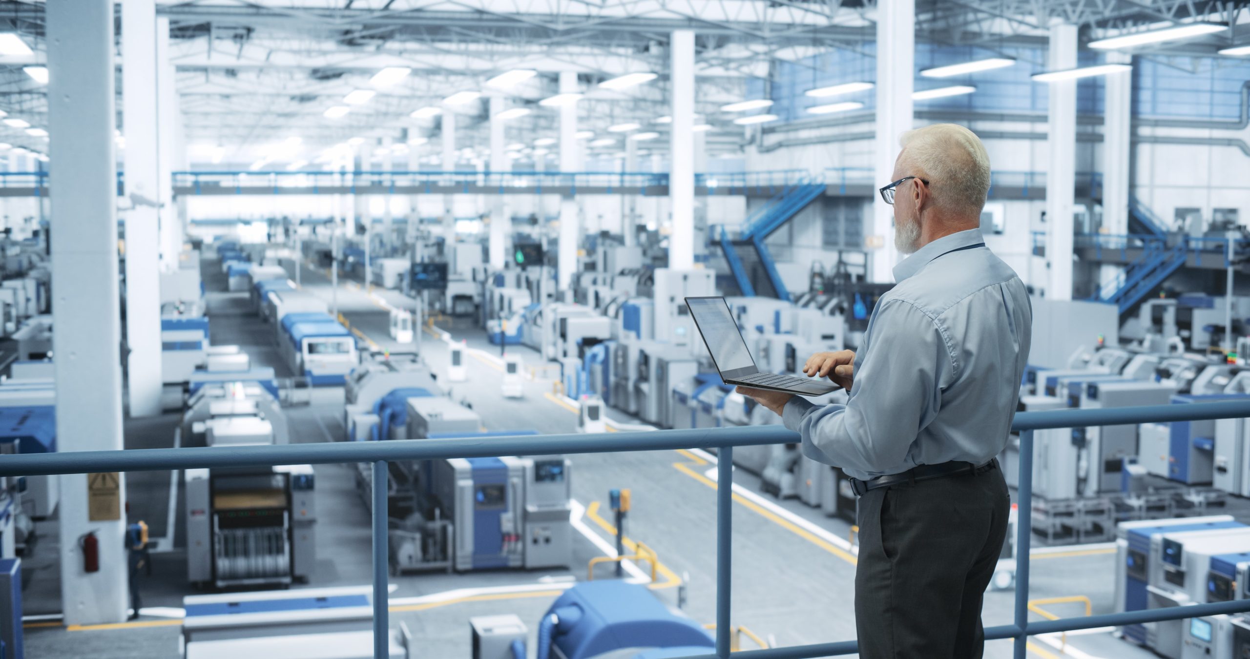 engineer looks at a production monitoring ystem on a laptop while looking out over a production floor